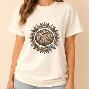 Può includere: Una t-shirt color crema con una grafica centrale raffigurante una pagnotta rotonda con la parte superiore incisa, posizionata sopra una lametta da pane. Il disegno è incorniciato da un motivo a raggiera stilizzato nei toni del marrone, nero e oro.