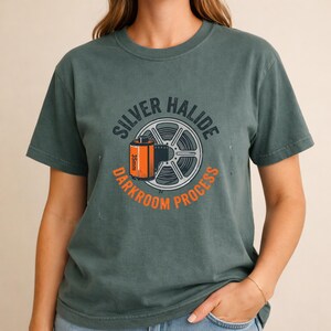 May include: A dark teal t-shirt featuring a graphic of a 35mm film canister and film reel. The text on the shirt reads "SILVER HALIDE DARKROOM PROCESS" in a circular design. The shirt is a solid color and has short sleeves.