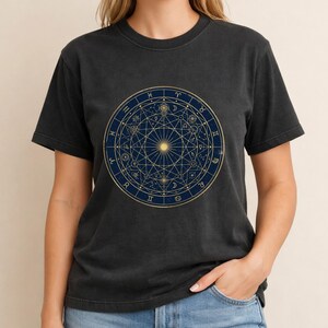 May include: A charcoal grey short-sleeve t-shirt featuring a large, circular graphic on the chest. The design is a dark blue celestial chart with gold lines, geometric patterns, and zodiac symbols arranged in a complex, symmetrical mandala style.