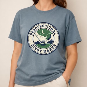 Może przedstawiać: Zakurzony niebieski t-shirt z krótkim rękawem z okrągłą grafiką o postarzanym wyglądzie. Wzór przedstawia kij golfowy uderzający w kawałek darni, półksiężyc oraz napis "PROFESSIONAL DIVOT MAKER" w ciemnoniebieskiej, okrągłej obwódce.