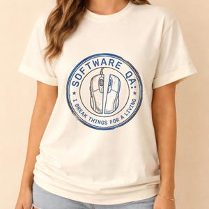 Puede incluir: Una camiseta de color crema con un gráfico circular azul en el pecho. El diseño incluye una ilustración de un ratón de ordenador partido por la mitad, rodeado por el texto "SOFTWARE QA: I BREAK THINGS FOR A LIVING" en una fuente negrita y desgastada.