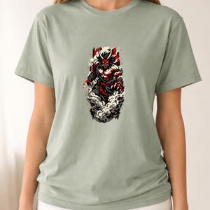 May include: Sage green t-shirt featuring a detailed graphic of a red and black demon-like figure emerging from a cloud of smoke. The illustration has a Japanese art style. The shirt is a short-sleeved crew neck.