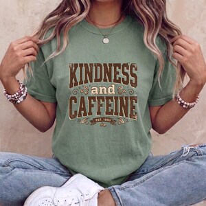 Puede incluir: Una camiseta de manga corta color verde salvia con texto marrón desgastado que dice KINDNESS and CAFFEINE y el texto más pequeño EST. 1993 debajo. El diseño incluye pequeñas ilustraciones gráficas de granos de café y hojas alrededor.
