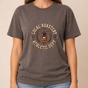 May include: Dark gray t-shirt featuring a circular graphic with the text "LOCAL ROASTERY ATHLETIC DEPT" surrounding a coffee grinder illustration. The graphic is in shades of brown, orange, and white. The shirt has short sleeves and a crew neck.