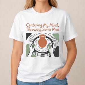 Può includere: Una t-shirt bianca con un'illustrazione grafica di un vaso in terracotta su un tornio con due mani che lo modellano. Sopra la grafica, il testo recita, Centering My Mind, Throwing Some Mud, in un carattere di colore marrone.
