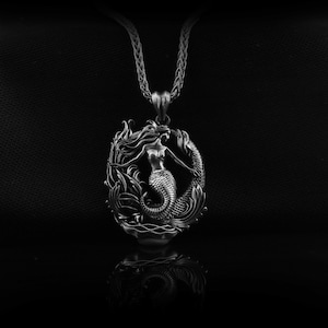 Pode incluir: Colar com pingente de prata com um design detalhado de sereia. O pingente mostra uma sereia com cabelo esvoaçante e cauda de peixe, rodeada por ondas e folhagens estilizadas. O colar tem uma corrente delicada.