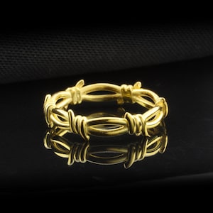 Puede incluir: Un anillo dorado con un diseño de alambre de púas. El anillo es circular y presenta múltiples púas a lo largo de su circunferencia. El anillo se muestra sobre una superficie negra reflectante.