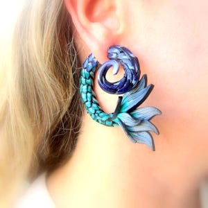 Könnte beinhalten: Ein Paar blau-türkisfarbene Ohrringe mit einem Meerjungfrauenschwanz-Design. Die Ohrringe haben eine spiralförmige Form mit einem strukturierten, schuppenartigen Muster in Türkis und Blau sowie einen fächerförmigen Schwanz.