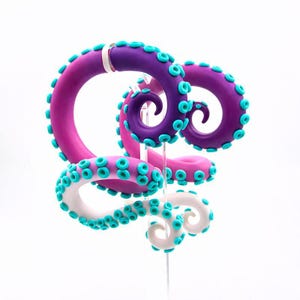 May include: A whimsical octopus tentacle sculpture in shades of purple, pink, and white, adorned with turquoise suction cups. The tentacles curl and twist, creating a dynamic and playful design. The sculpture is mounted on a clear stand.