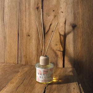 May include: A glass reed diffuser with a wooden top and reeds. The label reads "Snug Vanilla Blush Luxury Reed Diffuser" with pink accents. The diffuser is set on a wooden surface against a wooden backdrop.
