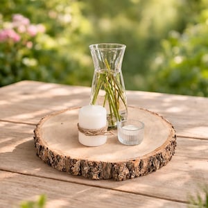 Op de afbeelding: Een rustieke houten schijf met een natuurlijke boomschorsrand houdt een heldere glazen vaas met groene stelen, een witte kaars vastgebonden met touw en een kleine glazen votief. De opstelling staat op een houten tafel.