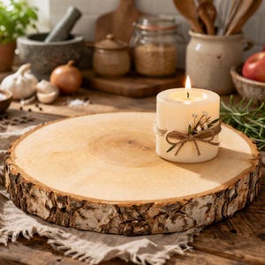 Op de afbeelding: Een brandende, ivoorkleurige pilaar kaars met een touw strik en bloem accenten staat op een ronde, natuurlijke houten schijf. De kaars is het middelpunt, met een rustieke, warme uitstraling. Andere keukenartikelen staan op de achtergrond.