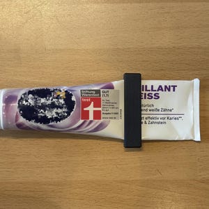 Könnte beinhalten: Eine Zahnpastatube mit weißem Verschluss und lila-weißem Design. Die Tube wird von einem schwarzen Clip gehalten. Der Text "ILLANT EISS" ist auf der Tube zu sehen.