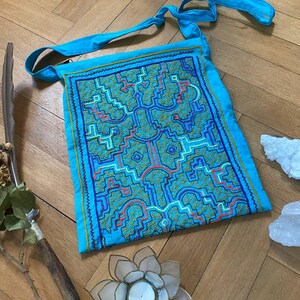May include: A turquoise shoulder bag with a geometric pattern in teal, orange, and blue. The bag has a long strap and is decorated with embroidered details. The bag is made of a soft fabric.