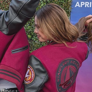 May include: A burgundy Starfleet Academy letterman jacket with black leather sleeves. The jacket features a Starfleet Academy patch on the sleeve and a Starfleet emblem on the back. The jacket has striped trim on the cuffs and waist.