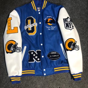 Puede incluir: Una chaqueta universitaria azul y blanca con detalles amarillos. La chaqueta presenta el logotipo de Los Angeles Rams, el logotipo de la NFL y el texto "Rams House Est. 1937". Las mangas son blancas con parches de casco de fútbol amarillo y negro.