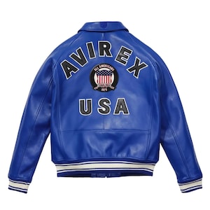 K&ouml;nnte beinhalten: Leuchtend blaue Lederjacke mit dem Wort "AVIREX" in wei&szlig;er Schrift auf dem R&uuml;cken. Darunter steht das Wort "USA", dar&uuml;ber ein kreisf&ouml;rmiger Aufn&auml;her mit der amerikanischen Flagge. Die Jacke hat einen klassischen Bomberstil mit wei&szlig;-blauen Streifen an den B&uuml;ndchen.