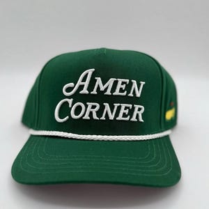 Può includere: Un cappellino da baseball verde scuro con la scritta "Amen Corner" in bianco. Il cappellino ha un dettaglio di corda bianca lungo il bordo e un piccolo logo sul lato.