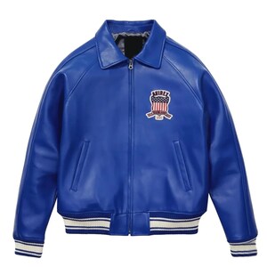 K&ouml;nnte beinhalten: Eine leuchtend blaue Lederjacke mit klassischem Kragen und Rei&szlig;verschluss. Die Jacke hat einen gerippten Saum und B&uuml;ndchen mit wei&szlig;en Streifen. Ein gestickter Aufn&auml;her mit dem Text "AVIREX" und einem patriotischen Design befindet sich auf der Brust.