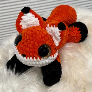 May include: A handmade orange, white, and black crocheted fox plush toy with large, glossy black eyes and gold glitter accents. The fox has a fluffy texture and is lying down, showcasing its detailed craftsmanship.