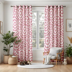 May include: Two pink and white patterned curtains hang in front of a window. The curtains feature a repeating pattern of white ovals on a pink background. A wooden chair with a grey cushion and blanket sits in front of the window. A cat and a small dog are on the floor.