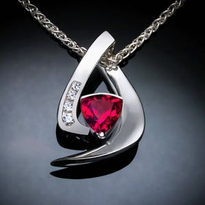 May include: A silver pendant necklace with a red triangular gemstone and smaller clear gemstones. The pendant is shaped like a stylized teardrop.