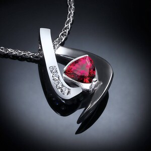 May include: A silver necklace with a red gemstone pendant. The pendant is shaped like a heart and has a small diamond on each side. The necklace is on a black background.