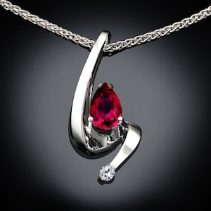 May include: A silver pendant necklace with a pear-shaped red gemstone and a smaller clear gemstone. The necklace has a braided chain.