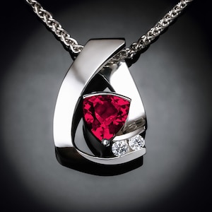 May include: A silver pendant necklace with a red gemstone and two smaller diamonds. The pendant is shaped like a teardrop and the gemstone is set in a triangular shape.