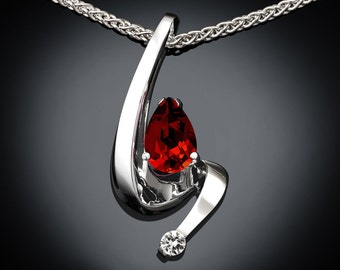Puede incluir: Un collar de plata con un colgante que tiene una piedra roja en forma de pera y una piedra transparente más pequeña. El colgante está en una cadena de plata.