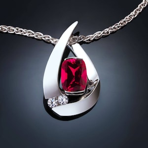 May include: A silver pendant necklace featuring a large, rectangular, red gemstone. The pendant has a teardrop design with two smaller, round, clear gemstones below the red stone. The chain is a silver, braided style.