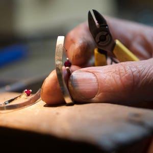 May include: A close-up of a person's hand holding a silver ring with a red gemstone. The ring is being worked on with a pair of small pliers.