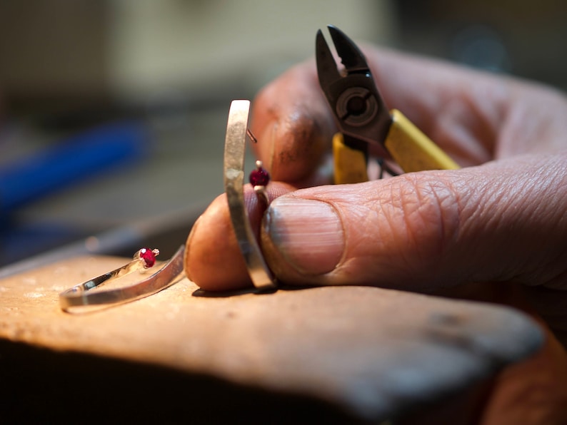 May include: A close-up of a person's hand holding a silver ring with a red gemstone. The ring is being worked on with a pair of small pliers.
