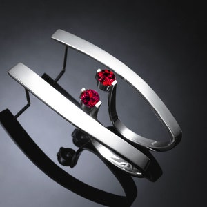 May include: A pair of silver earrings with a curved design and two red gemstones.