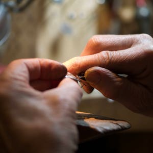 May include: Close-up of a person's hands working on a piece of jewelry with a small tool. The jewelry appears to be silver or white gold.