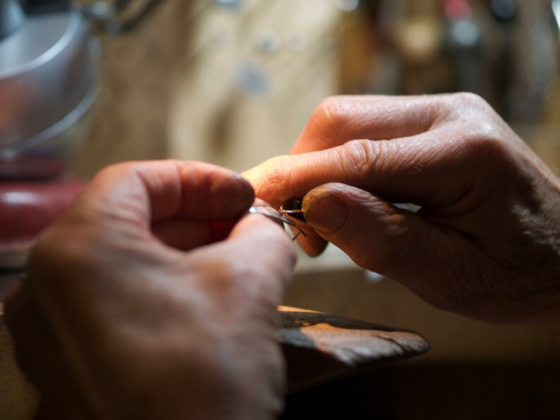 May include: Close-up of a person's hands working on a piece of jewelry with a small tool. The jewelry appears to be silver or white gold.