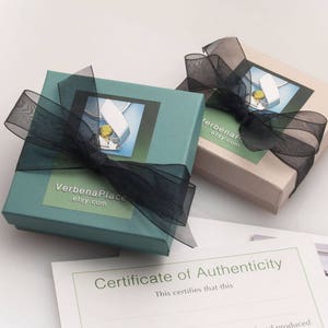 May include: Two teal and beige gift boxes tied with black ribbon. The boxes have a white label with the text "Verbena Place .etsy.com" and a picture of a silver pendant with a green gemstone. A certificate of authenticity is visible in the background.