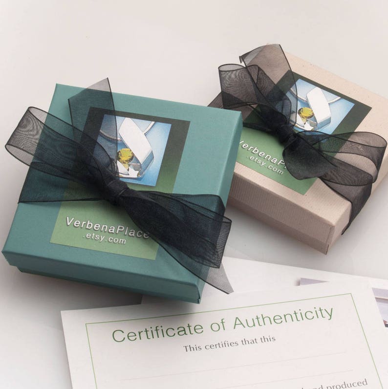May include: Two teal and beige gift boxes tied with black ribbon. The boxes have a white label with the text "Verbena Place .etsy.com" and a picture of a silver pendant with a green gemstone. A certificate of authenticity is visible in the background.