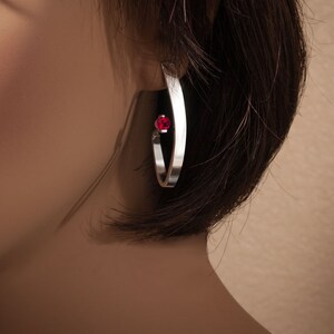 May include: Silver hoop earrings with a single red gemstone accent.