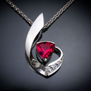 May include: A silver pendant necklace featuring a vibrant red triangular gemstone at its center, accented by three smaller white gemstones. The pendant has a flowing, abstract design and hangs from a silver chain.