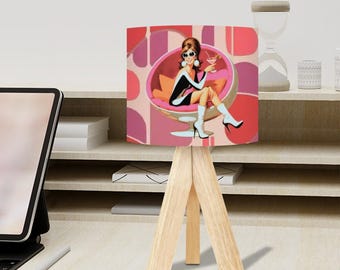 May include: A small wooden tripod lamp with a retro-style lampshade. The lampshade features a vintage illustration of a woman in a pink chair, holding a cocktail. The background is a pattern of pink and red shapes. The lamp is on a desk.