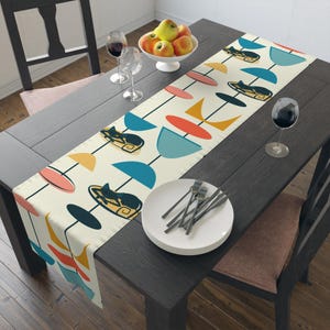May include: A cream-colored table runner with a mid-century modern design featuring black cat silhouettes, and abstract shapes in orange, yellow, and blue. The runner is placed on a dark wooden table, with plates, silverware, and wine glasses.