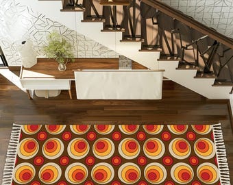 May include: A retro-style area rug with a brown background and repeating circles in red, orange, and yellow. The rug has a fringed edge and is placed on a hardwood floor. The rug is under a staircase with a wooden handrail.