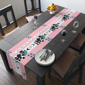 Könnte beinhalten: Ein rosa Tischläufer mit einem sich wiederholenden Muster aus schwarzen Pudeln mit blauen Hüten, weißen Streifen und türkisfarbenen Sternen. Der Läufer liegt auf einem dunklen Holztisch, der mit Tellern, Besteck und Weingläsern gedeckt ist.