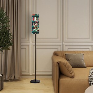 May include: A black floor lamp with a rectangular lampshade featuring a colorful illustration of a woman in a library. The lamp has a slender, black metal pole and a round base. The background includes a tan sofa and a wall.