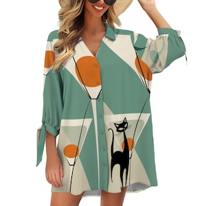 May include: A button-down shirt dress with a unique design. The dress features a teal, orange, and cream geometric pattern with a black cat illustration. The sleeves are three-quarter length with tie details. The dress has a V-neck collar and orange buttons.