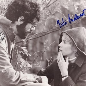 May include: A black and white photograph featuring two individuals, with the text Elliott Gould signed on the left and Bili Anderson signed on the right. The person on the left has curly hair, a beard, and glasses, while the person on the right wears a hooded coat.
