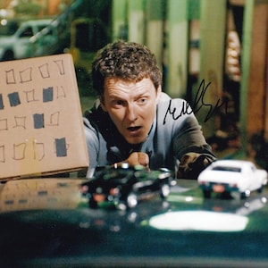 May include: A glossy photograph featuring a person with curly hair looking surprised, holding a cardboard sign with a grid of hand-drawn squares, some filled in with blue ink. A black signature is visible on the right side of the image.