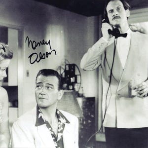 Peut inclure: Une photographie en noir et blanc montrant une femme aux cheveux ondulés, un homme portant une chemise à motifs et un homme en veste de costume tenant un combiné téléphonique. L'image porte la signature manuscrite Nancy Olson à l'encre noire.
