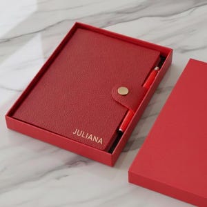 Könnte beinhalten: Ein rotes Leder-Notizbuch und Stift-Set in einer passenden Box. Das Notizbuch hat einen goldenen Knopfverschluss und den Namen "JULIANA" in goldfarbenen Buchstaben. Der Stift ist rot und passt in eine Schlaufe an der Seite des Notizbuchs.
